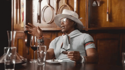 Man in cowboy hat gesturing at table with glasses and decanter.